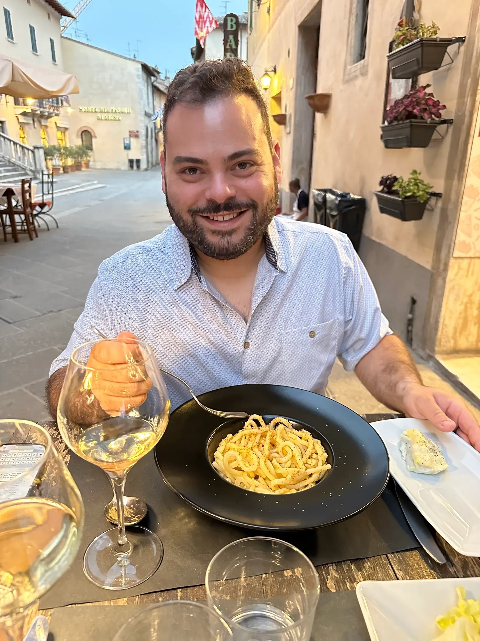 Me at an outdoor table in a Tuscan town with a plate of pici and a glass of white wine